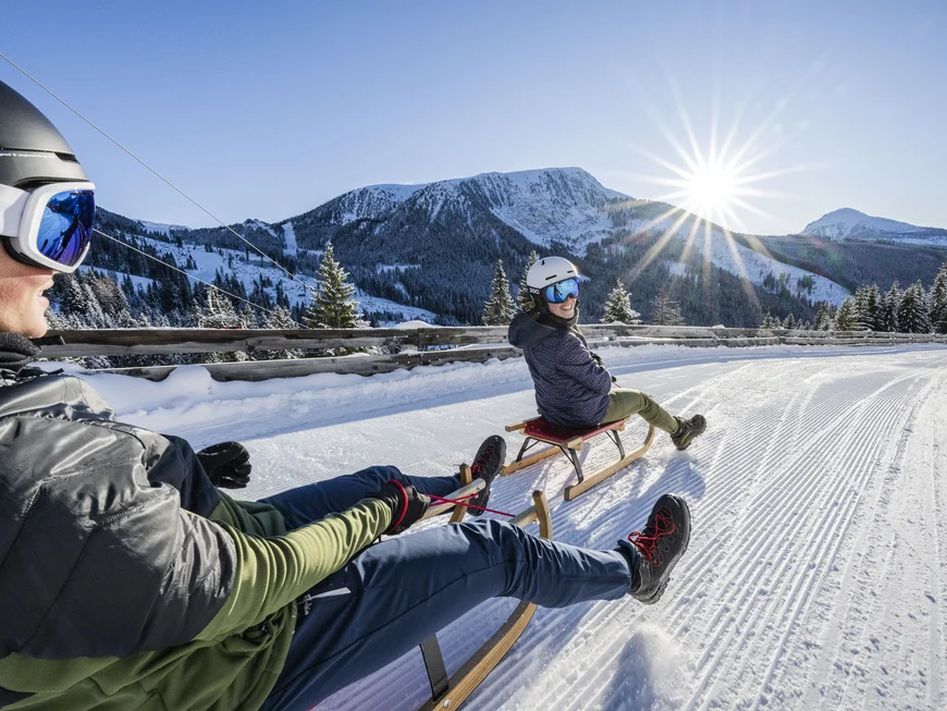 Ihr werdet dieses Skihotel in den Dolomiten lieben. Zwei Personen beim Rodeln auf einer Schneepiste vor Bergpanorama bei Sonnenschein