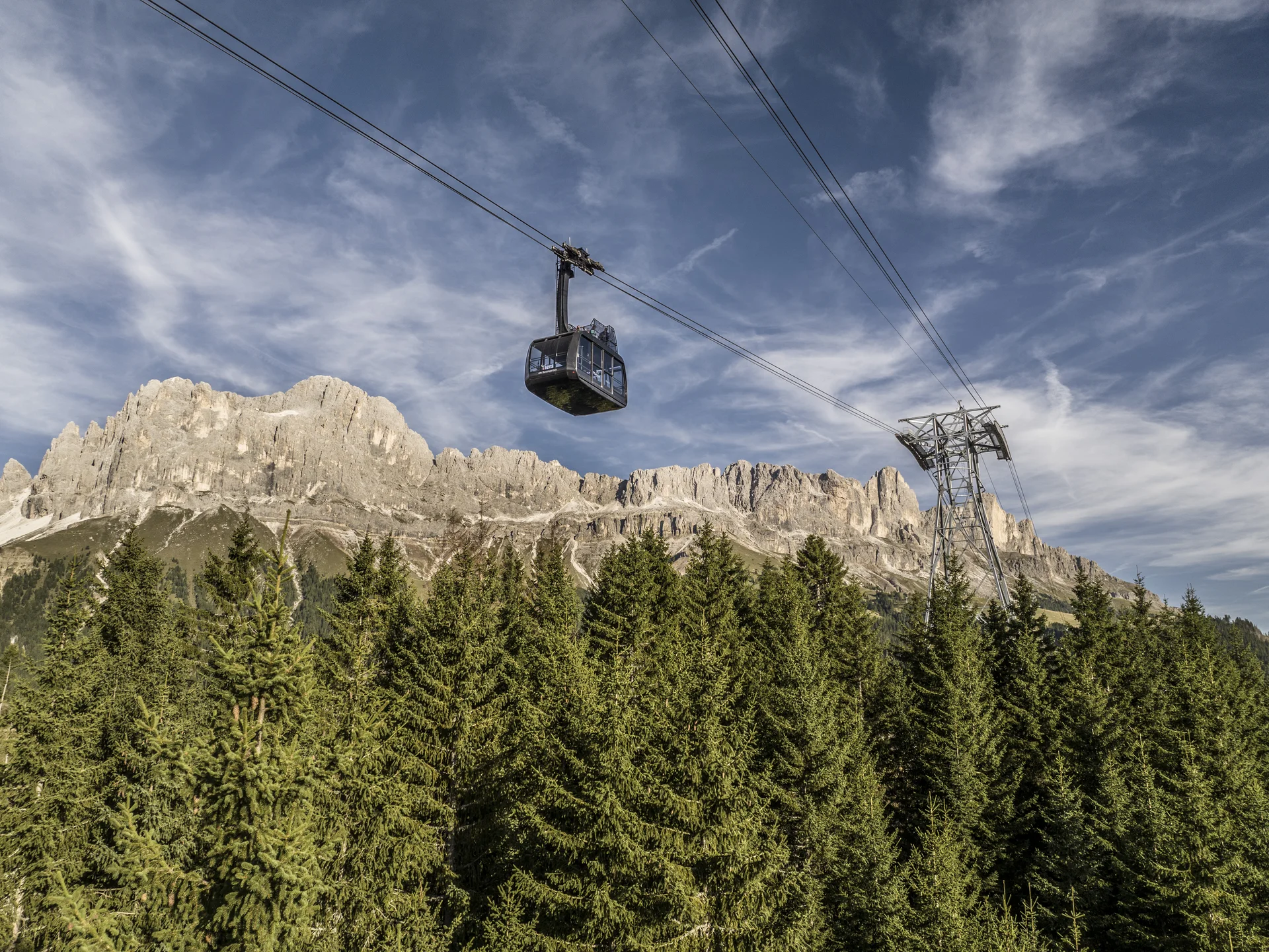 Seid ihr bereit für euren Sommerurlaub in den Dolomiten? Seilbahn über Tannenwald mit Bergkulisse und bewölktem Himmel