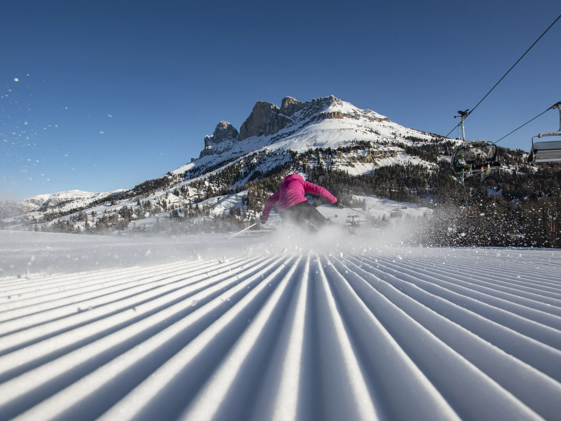 Ihr werdet dieses Skihotel in den Dolomiten lieben. Skifahrer auf frisch präparierter Piste vor schneebedecktem Berg und Sessellift