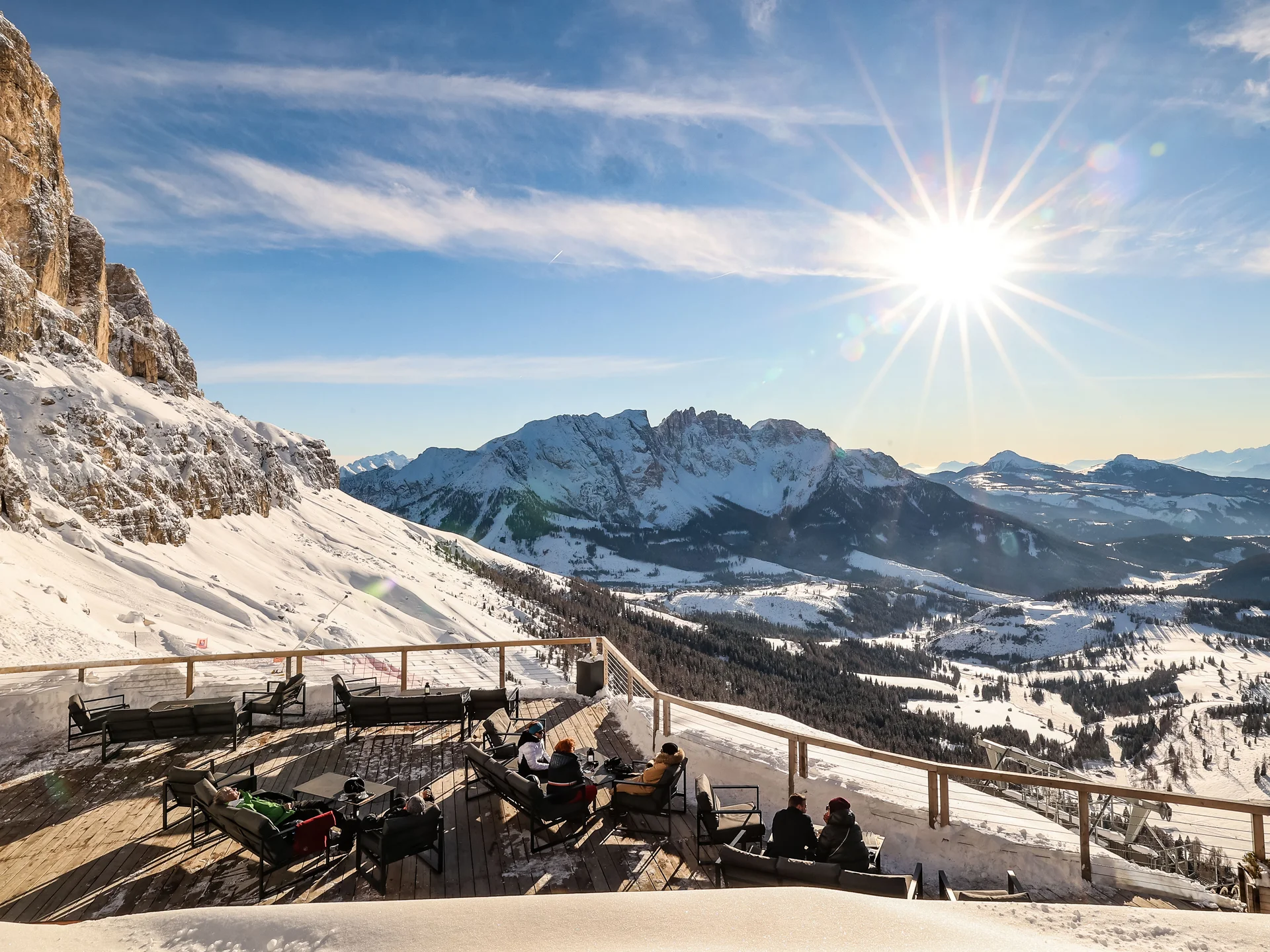 Ihr werdet dieses Skihotel in den Dolomiten lieben. Sonnige Bergterrasse mit Menschen und schneebedeckten Gipfeln im Hintergrund